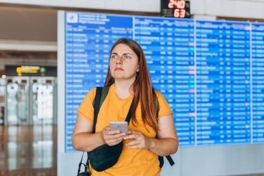 30 'lu yaşlarda cep telefonunu elinde tutan kafası karışmış bir kadın. İnternet' ten bilgi panosunun yakınındaki Trip Destination 'ı kontrol ediyor. Gecikmeli uçuş. Şok edici derecede kötü haberler alıyor.