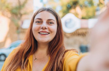 Şehirde yürürken selfie çeken gülümseyen genç bir kadın. Sosyal medya uygulamasında paylaşmak için dışarıda poz veren neşeli kız öğrenci. Beyaz kadın.