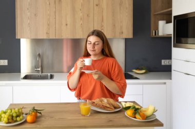 Dertsiz, mutlu, güler yüzlü bir kadın. Tembel bir sabahta evde kahve içiyor. Genç bayan modern mutfakta taze aromatik kahvenin tadını çıkarıyor.