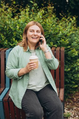 Moda kıyafetleri içindeki mutlu kızıl kadın telefonda bir fincan çayla konuşuyor. Modelleme. Güzel, şık bir kız akıllı telefondan arıyor ve açık hava bankında oturuyor.