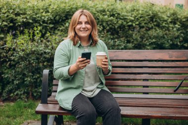 Güzel, gülümseyen 30 'lu yaşlarda, elinde kahve fincanı olan, akıllı telefon kullanan ve dışarıda duran bir kadın. Telefon iletişimi. Şehir parkında yürüyen neşeli genç bir kadın, şehir yaşam tarzı konsepti..