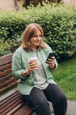 Güzel, gülümseyen 30 'lu yaşlarda, elinde kahve fincanı olan, akıllı telefon kullanan ve dışarıda duran bir kadın. Telefon iletişimi. Şehir parkında yürüyen neşeli genç bir kadın, şehir yaşam tarzı konsepti..
