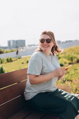 Sıradan giyinmiş 30 'lu yaşlardaki neşeli kadın bankta oturuyor ve elinde şehir sokaklarında iletişim kurmak için akıllı telefon aleti tutuyor. Kadın telefonda konuşuyor ve dışarıda dinleniyor..