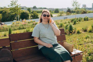 Sıradan giyinmiş 30 'lu yaşlardaki neşeli kadın bankta oturuyor ve şehir caddesinde akıllı telefondan konuşuyor. Dişi dışarıda dinleniyor. Şehir konsepti
