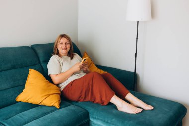 Akıllı telefon tutan genç bir kadın, cep telefonu modern teknolojisini kullanıyor, cep telefonu uygulamalarını kontrol ediyor, mesajlaşıyor, video izliyor, internetten alışveriş yapıyor. Evdeki kanepede oturan kız. Tam boy gövde