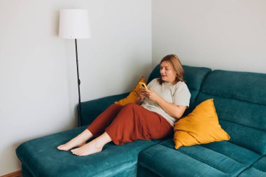 Akıllı telefon tutan genç bir kadın, cep telefonu modern teknolojisini kullanıyor, cep telefonu uygulamalarını kontrol ediyor, mesajlaşıyor, video izliyor, internetten alışveriş yapıyor. Evdeki kanepede oturan kız. Tam boy gövde