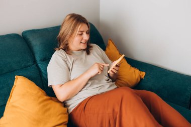 Akıllı telefon tutan genç bir kadın, cep telefonu modern teknolojisini kullanıyor, cep telefonu uygulamalarını kontrol ediyor, mesajlaşıyor, video izliyor, internetten alışveriş yapıyor. Evdeki kanepede oturan kız..