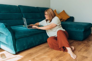 Gözlüklü güzel bir kadın evdeki kanepenin yanındaki defterde çalışıyor. Eğitim, iş ve konfor kavramları. İçerde çalışan 30 'lu yaşlarda bir kadın.