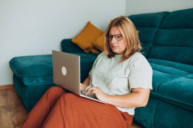 Üzgün, şüpheci, gözlüklü ciddi bir kadın evdeki kanepenin yanındaki defterde çalışıyor. Eğitim, iş ve konfor kavramları. Olumsuz duygular, kanepede oturan düşünceli kız.