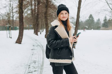 Güzel bir kadın parkta yürüyor, telefonu internetten alışveriş için kullanıyor, kışın karlı bir günde.