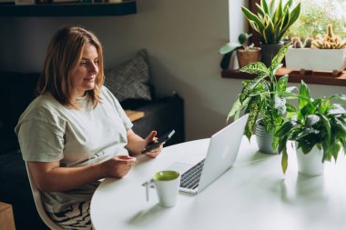 30 'lu yaşlardaki mutlu bir kadın bilgisayarında kredi kartı ve telefonuyla ev ofisinin masasında alışveriş yapıyor. Genç beyaz bir kadın bilgisayarından internetten alıyor..