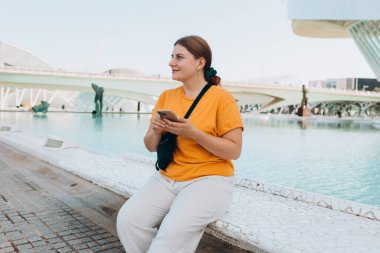 Dışarıda telefonla dolaşan genç bir kadın. Seyahat, turizm ve tatil kavramı. Ciudad de las Artes y las Ciencias de Valencia. Valencia Sanat ve Bilim Şehri..