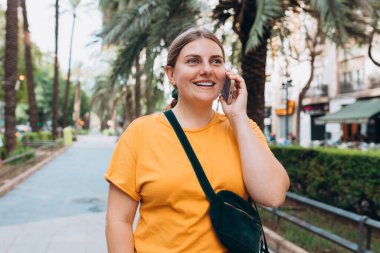 Parkta telefonla konuşan gülümseyen güzel bir kadının portresi. Neşeli kadın turist, sıradan giyinmiş, dışarıda dinleniyor.