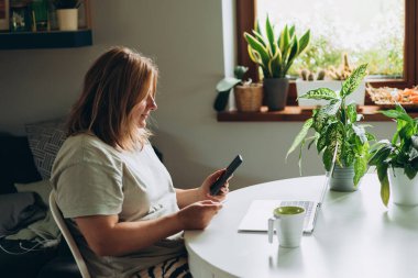 30 'lu yaşlardaki mutlu bir kadın bilgisayarında kredi kartı ve telefonuyla ev ofisinin masasında alışveriş yapıyor. Genç beyaz bir kadın bilgisayarından internetten alıyor..