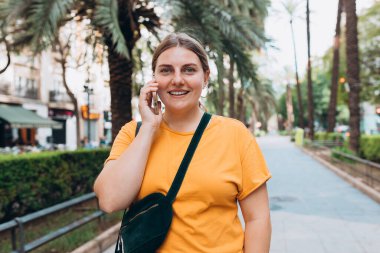 Parkta telefonla konuşan gülümseyen güzel bir kadının portresi. Neşeli kadın turist, sıradan giyinmiş, dışarıda dinleniyor.