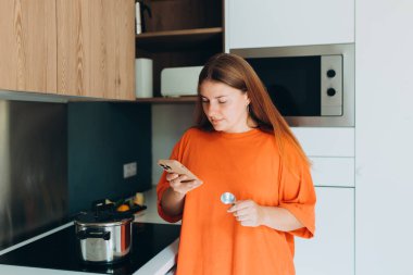 Mutfakta lezzetli yemekler yiyen bir kadın, ocağın yanında duran bir kız. Ev hayatı afişi. Yüksek kalite fotoğraf. Elinde kaşık ve telefon tutan heyecanlı genç bir kadının portresi.