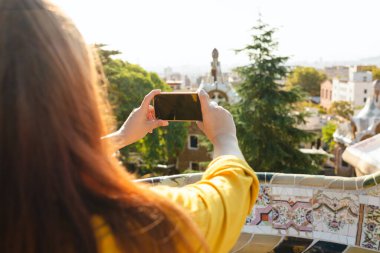 Kızıl saçlı kadın Guell Park 'tan Barselona' nın fotoğrafını çekiyor. Seyahat, turizm, teknoloji ve tatil kavramı. Arka görünüm.