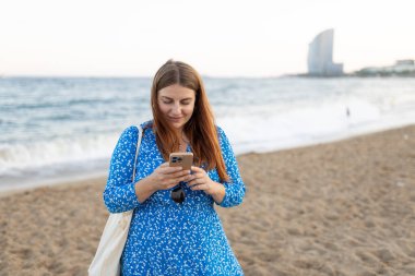 30 'lu yaşlarda, güzel, kızıl saçlı, mavi elbiseli, kumsalda akıllı telefon kullanan bir kadın. Seyahat, turizm ve tatil kavramı