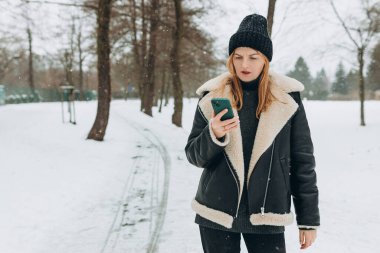 Kızgın kadın dışarıda hayal kırıklığıyla akıllı telefonuna bakıyor. Güzel bir kadın kışın karlı bir günde parkta yürüyor.
