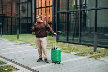 Sıradan giyinmiş 30 'lu yaşlardaki neşeli turist şehir sokaklarında iletişim kurmak için elinde akıllı telefon aleti tutuyor. Yeşil bavullu mutlu adam otelin yakınında telefonla konuşuyor..