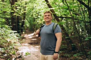 Turist sırt çantasıyla gülümseyen 30 'lu yaşlardaki adam bir ormanda duruyor. Yürüyüş, aktif yaşam tarzı, macera ve turizm, insanların duyguları