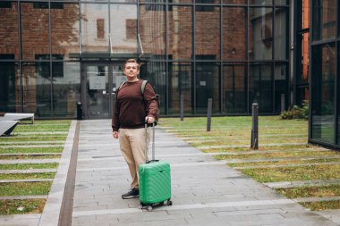 Yeşil bavulu dışarı çıkaran 30 'lu yaşlardaki mutlu adam. Genç adam seyahat çantasıyla sokakta yürüyor. Otelin yakınında bagajını taşıyan bir erkek. Seyahat Moda