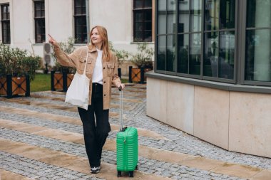 Yeşil valizlerle şehir caddesinde yürüyen günlük kıyafetli neşeli genç bir kadın. 30 'lu yaşlarda, bavullu bir kadın şehir geçmişini işaret ediyor. Seyahat Moda.