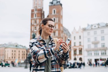Güz günü Krakow 'da pazar meydanında yürüyen ve cep telefonu tutan gözlüklü güzel bir kadın. Telefon iletişimi. Şehir yaşam tarzı konsepti. Sosyal ağları kontrol et