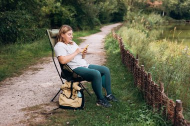 Göl kenarındaki güvertede oturan ve elinde cep telefonu olan rahat mutlu kadın yeni bir seyahat planlıyor. Kamp, turizm, teknoloji ve seyahat konsepti. 30 'lu yaşlarda genç bir kadın ormanı keşfediyor.