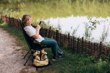 Göl kenarındaki güvertede oturan ve elinde cep telefonu olan rahat mutlu kadın yeni bir seyahat planlıyor. Kamp, turizm, teknoloji ve seyahat konsepti. 30 'lu yaşlarda genç bir kadın ormanı keşfediyor.