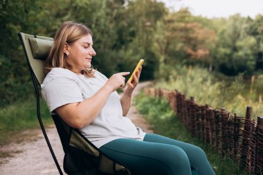 Göl kenarındaki güvertede oturan ve elinde cep telefonu olan rahat mutlu kadın yeni bir seyahat planlıyor. Kamp, turizm, teknoloji ve seyahat konsepti. 30 'lu yaşlarda genç bir kadın ormanı keşfediyor.