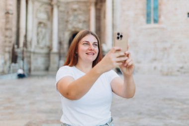 Mutlu kızıl kadın kentin arka planında selfie çekiyor. Teknoloji kavramını kullan, yazın Avrupa 'yı gezmek. Çekici bayan turist yeni şehri keşfediyor.