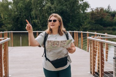 Güneş gözlüklü şık hippi kadın elinde kağıt harita, işaret ediyor, sırt çantası takıyor ve doğada dinleniyor. Seyahat ve seyahat tutkusu konsepti. İnanılmaz soğuk bir an.