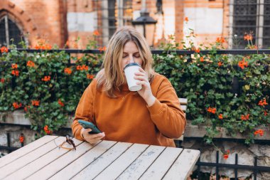 Elinde biyo-kahve fincanı ve akıllı telefon tutan kadın şehrin caddesindeki kafe terasında oturuyor. Şehir konsepti. Mutlu kız kahve masasında oturuyor ve cep telefonu kullanıyor.