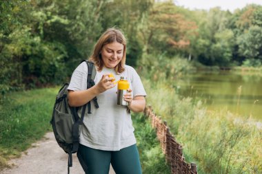 Happy smiling 30s women with backpack is holding thermos mug. Hot tea or other beverage on summer day. Lifestyle adventure concept. Woman drink water from the bottle clipart