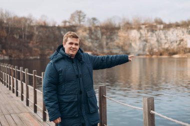 Seyahat, aktif yaşam tarzı, insanlar ve hobi konsepti. Sarışın erkek, sonbahar zamanı, dışarıyı işaret ediyor. Gezgin eko-turizmin tadını çıkarıyor. Göl kıyısındaki tahta iskelede duran mutlu adam