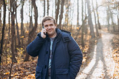Mutlu 30 'lu yaşlardaki genç adam telefonla konuşur, dinler ve ormanda yürür. Telefon iletişimi. Genç erkek doğa yürüyüşü yapıyor..
