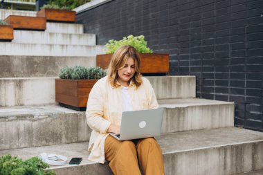 Merdivenlerde oturup laptopla çalışan mutlu sarışın kadın, hızlı bağlantı. Dizüstü bilgisayarı olan güzel bir kız sokakta oturuyor. Serbest çalışan biri. Uzaktan çalış.