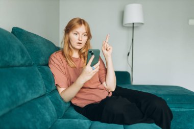 Akıllı telefonlu gülümseyen genç bir kadın işaret parmağıyla işaret ederek evdeki koltukta oturmak harika bir fikir. Güzel 30 'lu yaşlarda cep telefonuyla konuşan güzel bir kız.