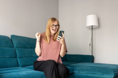 Heyecanlı bir kadın cep telefonunu evdeki koltukta tutarak başarıyı kutluyor.