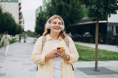 Şehir arka planında kablosuz kulaklıkla müzik dinleyen sevimli bir genç kızın yakın plan portresi. Müzik aşığı müziğin tadını çıkarıyor. Cep telefonu diktafonundan kadın sesi kaydı