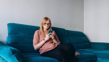 Üzgün, kuşkucu, ciddi bir kadın içeride iletişim kurmak için akıllı telefon aleti tutuyor. Negatif insanların duyguları, evdeki kanepede oturan düşünceli kız. 30 'lu yaşlarda elinde akıllı telefon tutan bir kadın..