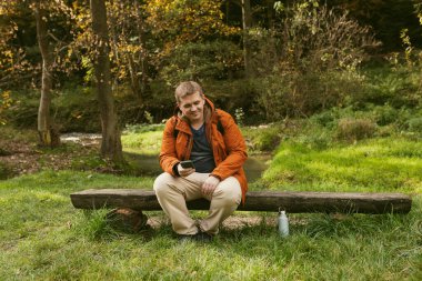 Sırt çantalı şık bir adam akıllı bir telefon tutuyor ve doğada dinleniyor. Tam vücut fotoğrafı. Aktif erkek yaşam tarzını dışarıda ağaç gövdesinde oturarak sürdürüyor. Yüksek kalite fotoğraf. Maceralar