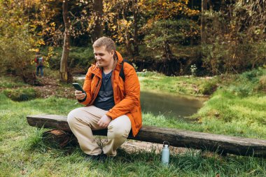 Sırt çantalı şık bir adam akıllı bir telefon tutuyor ve doğada dinleniyor. Tam vücut fotoğrafı. Aktif erkek yaşam tarzını dışarıda ağaç gövdesinde oturarak sürdürüyor. Yüksek kalite fotoğraf. Maceralar
