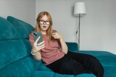 Akıllı telefonlu gülümseyen genç bir kadın işaret parmağıyla işaret ederek evdeki koltukta oturmak harika bir fikir. Güzel 30 'lu yaşlarda cep telefonuyla konuşan güzel bir kız.