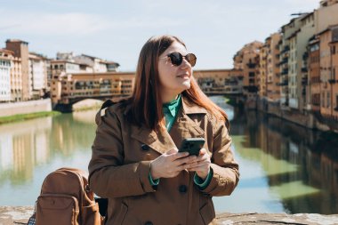 Floransa 'daki ünlü Old Bridge' de akıllı telefon kullanan 30 'lu yaşlardaki mutlu kız. İtalyan simgelerini ziyaret eden şık bir kadın. Seyahat, turizm ve tatil kavramı. Şehir caddesinde yürüyen kadın telefonu kontrol ediyor.