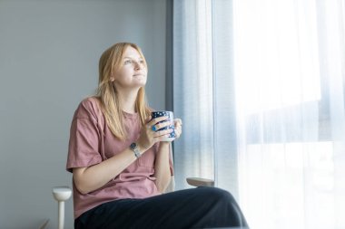 Neşeli, rüya gibi bir kadın elinde bir kupa sıcak içecekle, derin iyi düşüncelerle yüzünü çeviriyor, gülümsüyor, hayal kuruyor, aromatik bitki çayının tadını çıkarıyor.