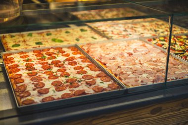 Restoran vitrininde otantik İtalyan pizzası. Cam vitrinli çeşitli pizzacılar. Salamlı pizzaya odaklan. Restorandaki pizza dilimlerinin görüntüsü.