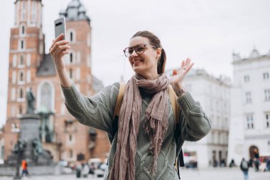 Gözlüklü mutlu kız kentin arka planında selfie çekiyor. Teknoloji kavramını kullan, sonbaharda Polonya 'yı gezmek. Krakow 'daki Pazar Meydanı, En İyi Turist Çekimi.