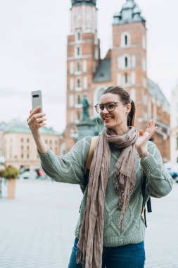 Gözlüklü mutlu kız kentin arka planında selfie çekiyor. Teknoloji kavramını kullan, sonbaharda Polonya 'yı gezmek. Krakow 'daki Pazar Meydanı, En İyi Turist Çekimi.